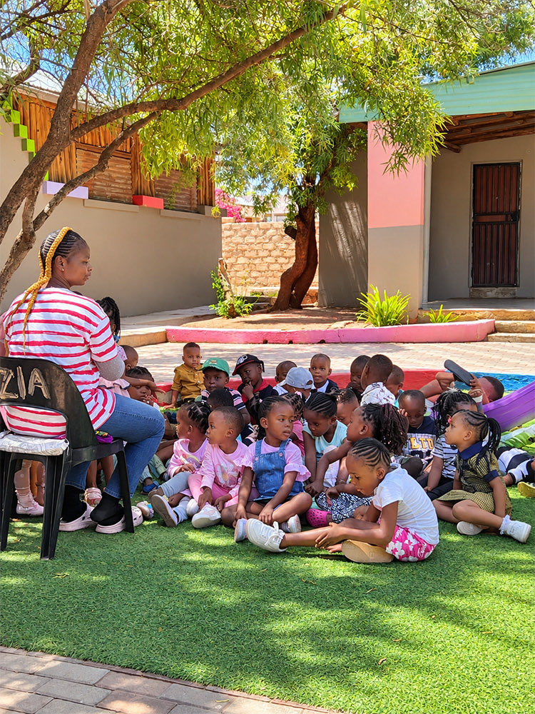 A teacher sitting with a large group of children on the grass under a tree