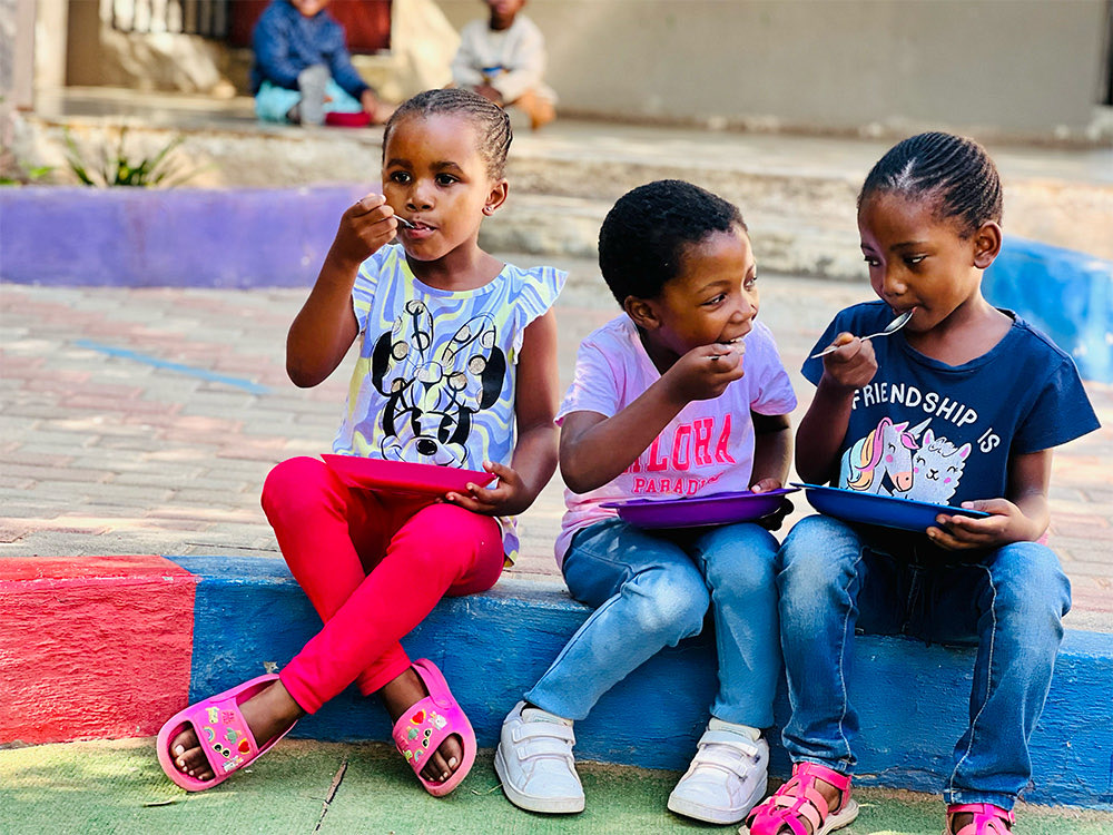 Three children sitting on a step eating lunch from coloured plates