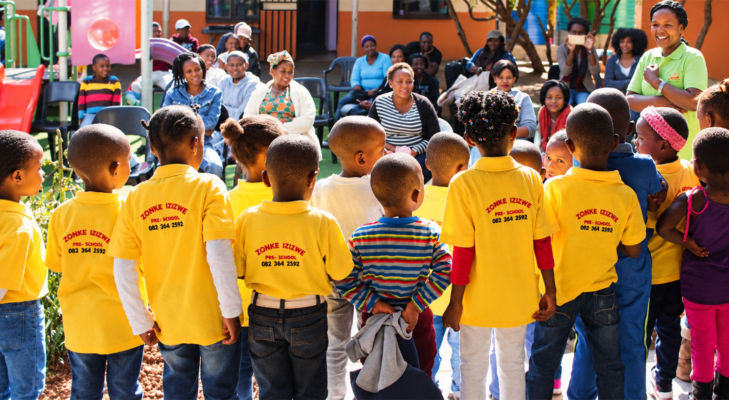 Zonke Izizwe pupils in yellow school shirts gathered in front of seated parents at the school