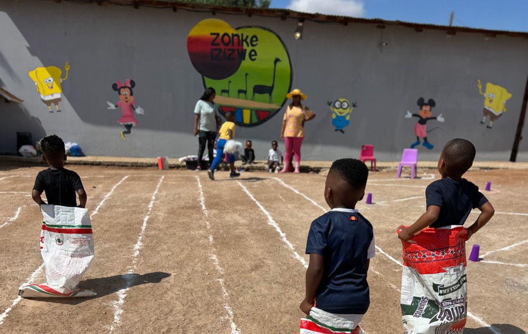 Children lining up for a sack race in front of the painted Zonke Izizwe mural