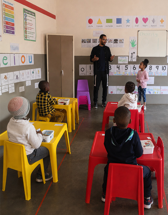 A teacher leading a lesson at Zonke Izizwe, children seated at yellow and red tables beneath alphabet and number charts